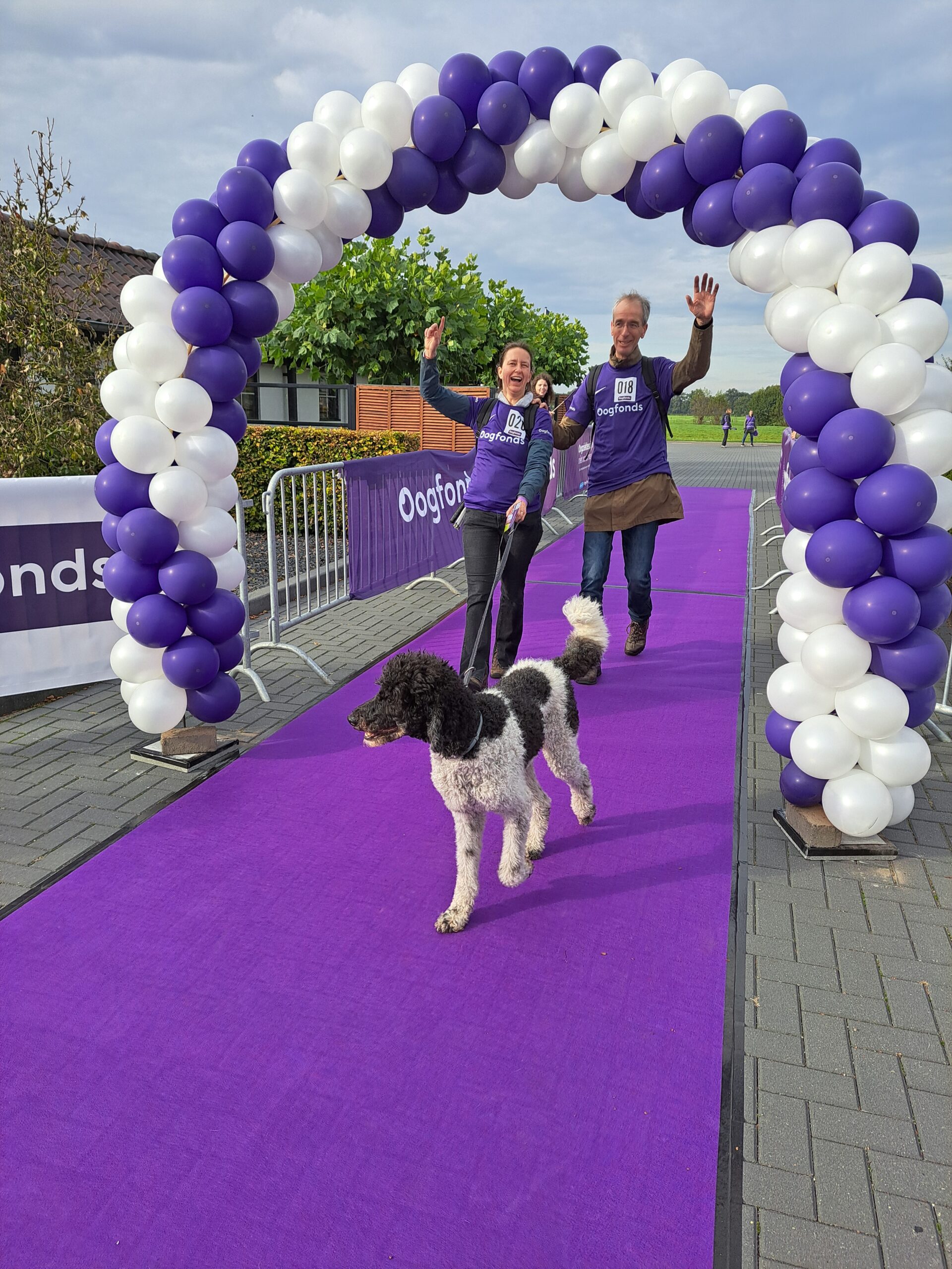 Twee mensen en een hulphond lopen juichend onder boog van ballonnen door bij Oogfonds Walk & ride