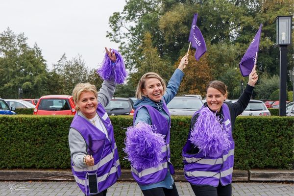 Drie enthousiaste medewerkers van het Oogfonds zwaaien met paarse vlaggen en pompons tijdens een actie voor een feestelijke inzamelingsactie.