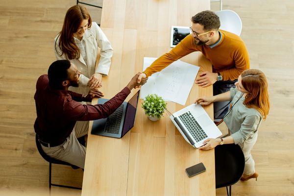 Vier collega’s overleggen rond een houten tafel; twee van hen schudden elkaar de hand, terwijl laptops, documenten, een plant en een telefoon op tafel liggen. Select 75 more words to run Humanizer.